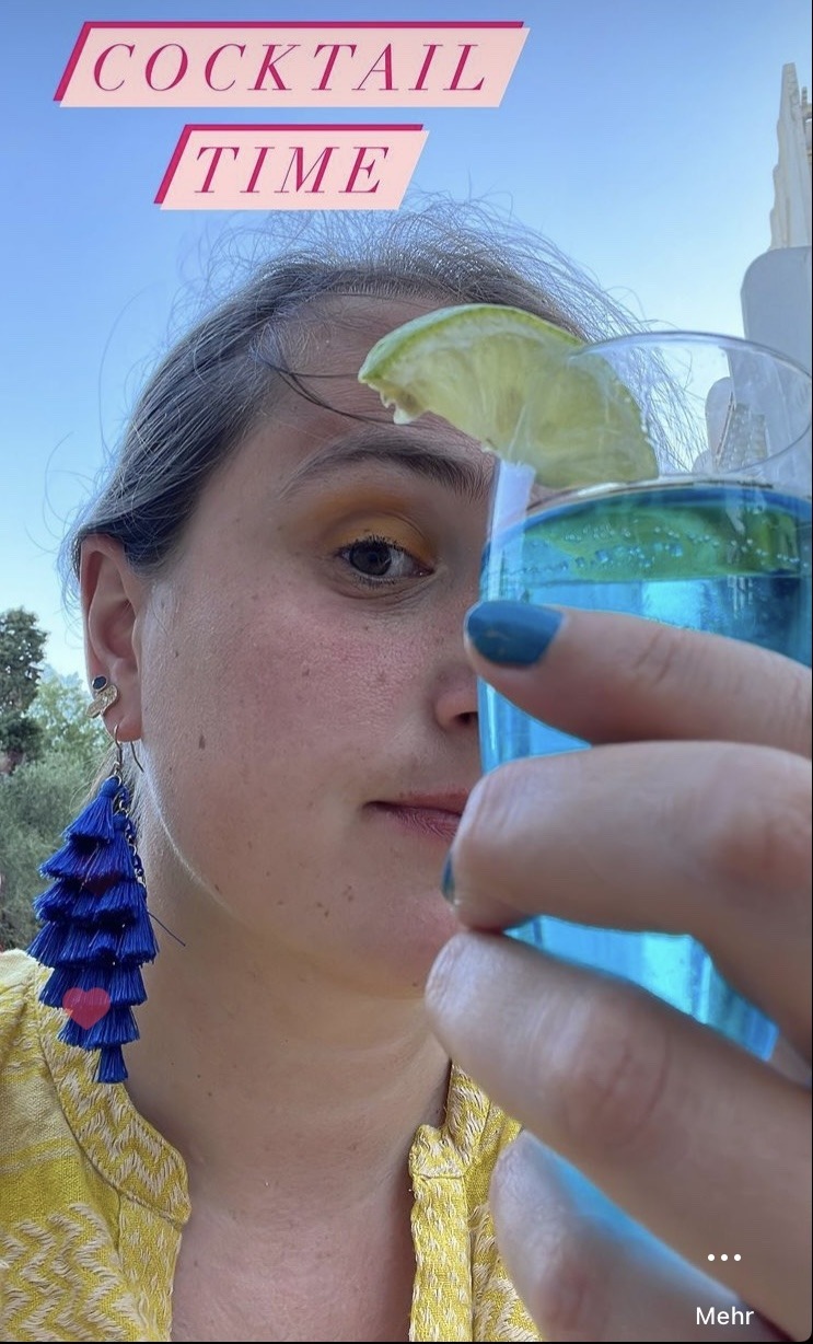 Frau mit großen blauen Ohrringen hält Cocktail Glas mit blauer Flüssigkeit vor linke Gesichtshälfte.