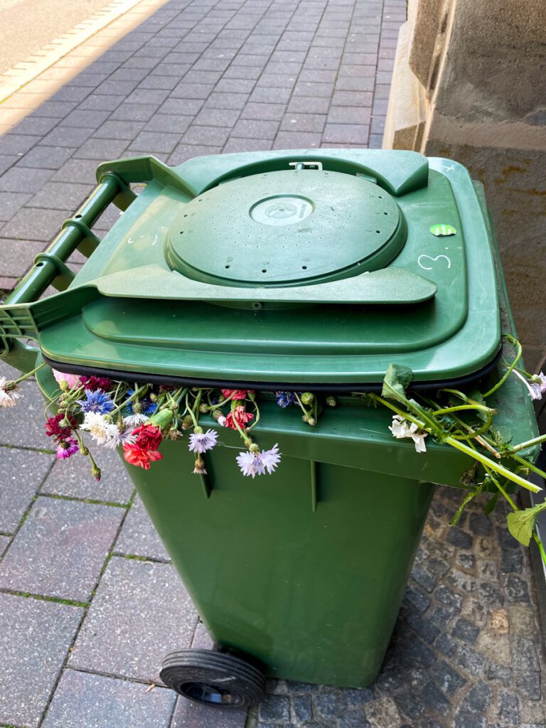 Blumen, die aus einer grünen Mülltonne heraushängen.